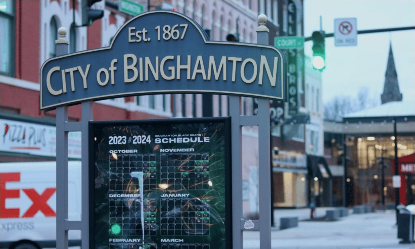 A downtown street with storefronts beneath a City of Binghamton sign.