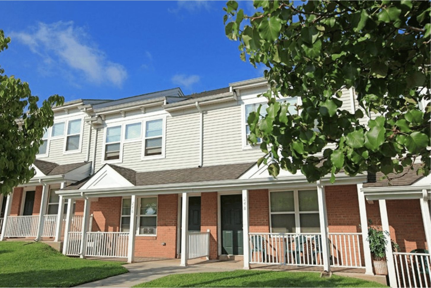 A row of two-story brick townhomes with covered porches behind a large shade tree.