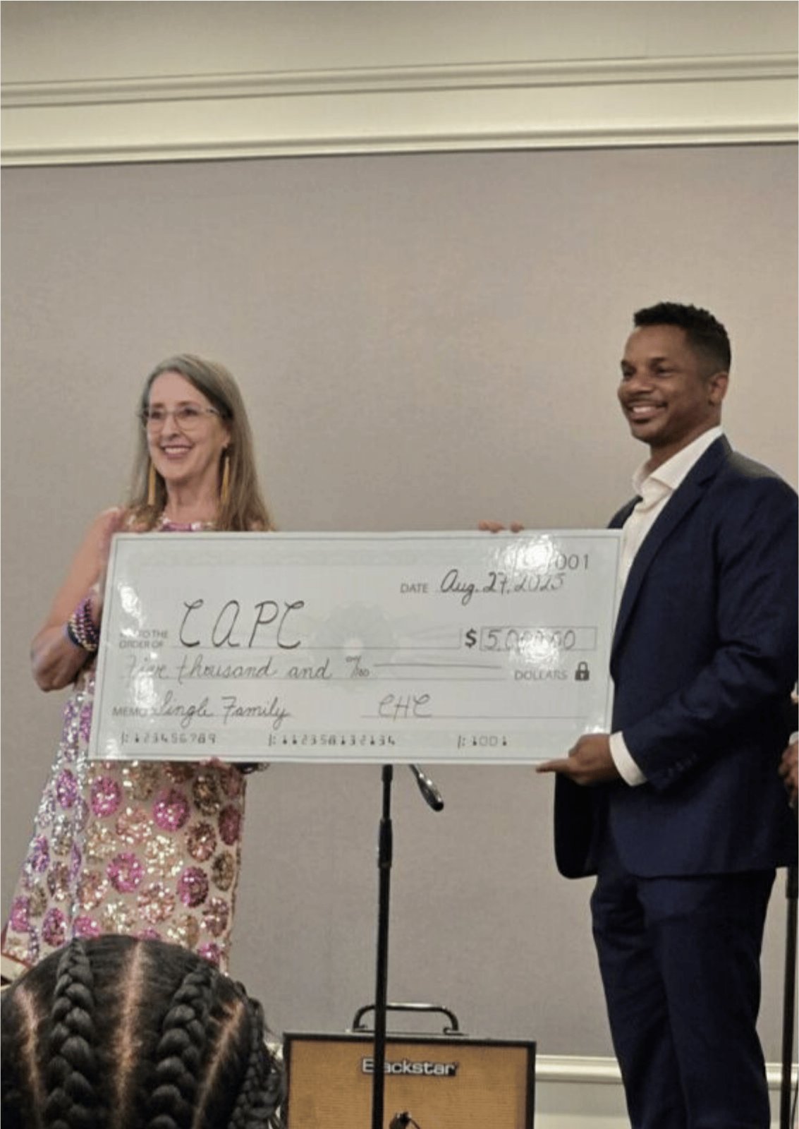 Two people holding an oversized ceremonial check marked CAPC at an indoor event.