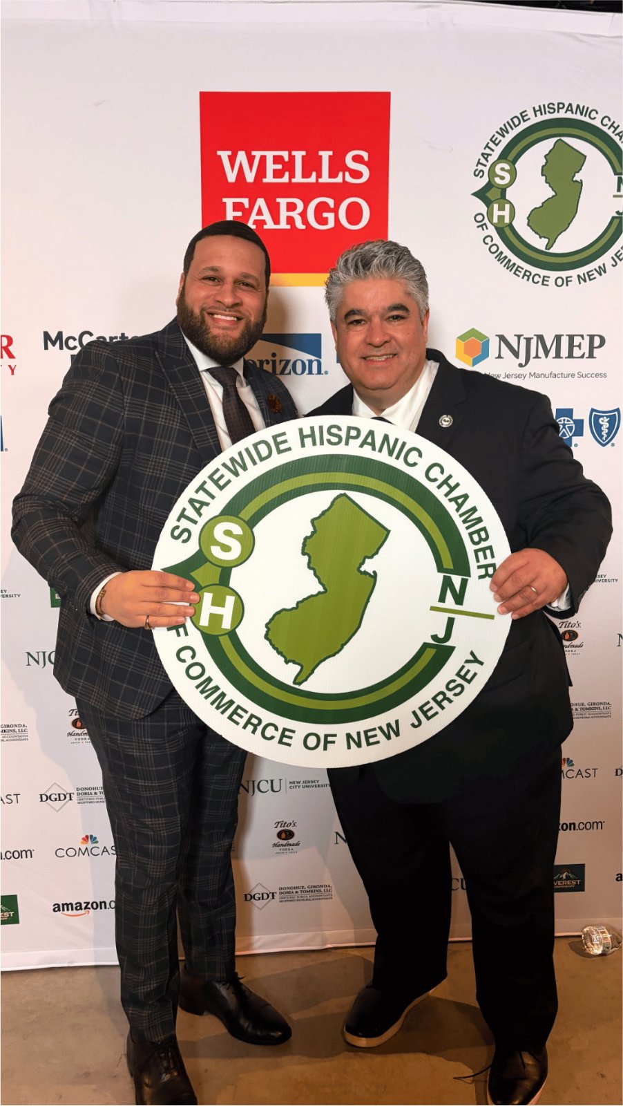 Two men standing together holding a Statewide Hispanic Chamber of Commerce of New Jersey sign at an event.