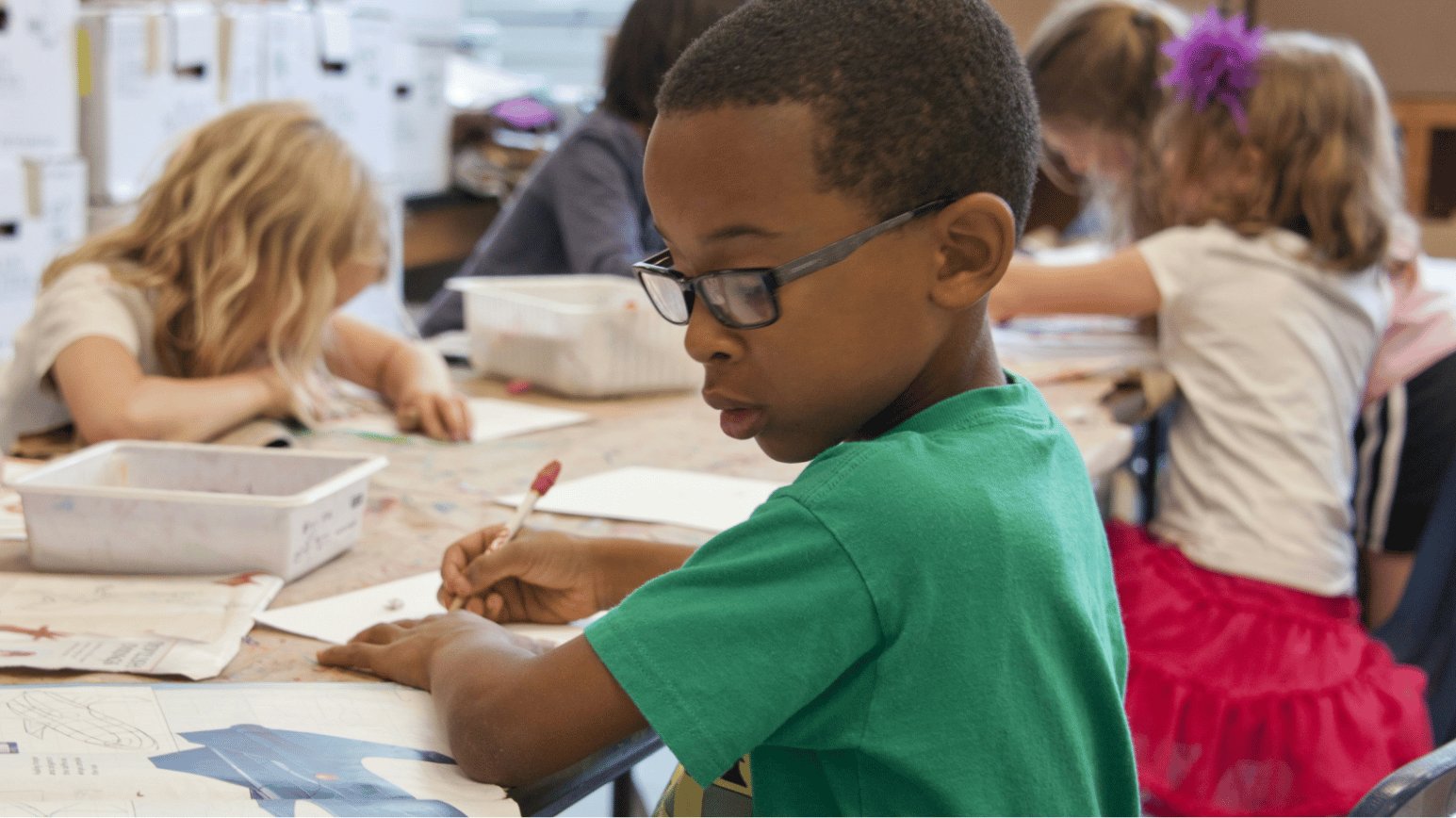 Young students working at a classroom table with a teacher nearby.